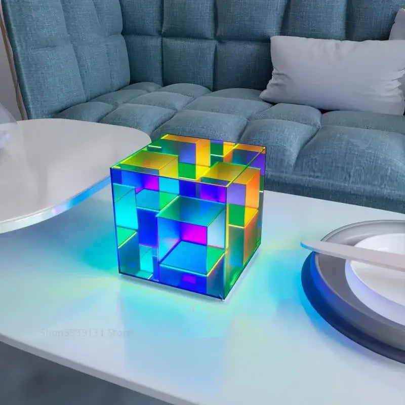 A translucent rainbow-colored decorbites led magic acrylic cube table lamp with geometric design on a white table