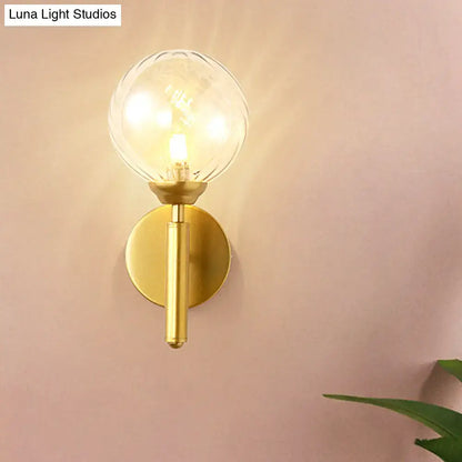 Modern brass wall lamp fixture with gold-toned cylindrical stem and clear textured glass globe for bedroom use
