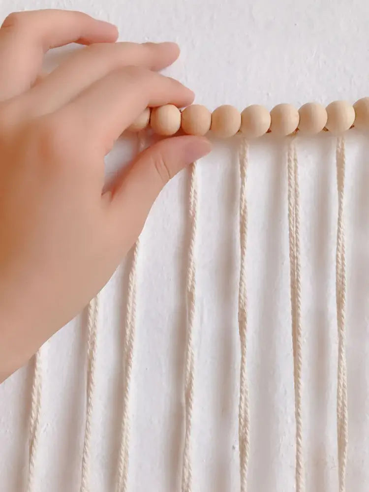 A hand gently holds light beige wooden beads on cream macrame cords in moroccan bohemian wall decor tapestry