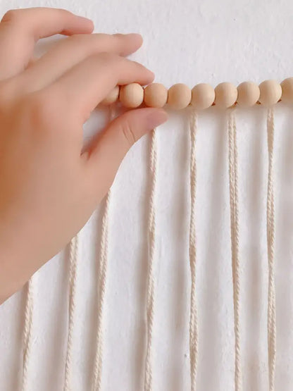 A hand gently holds light beige wooden beads on cream macrame cords in moroccan bohemian wall decor tapestry