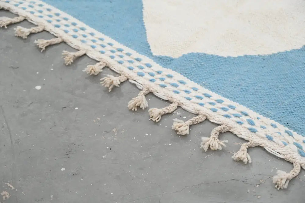 A cream-colored macrame fringe with woven pattern borders the edge of a blue and white moroccan handmade traditional azilal rug