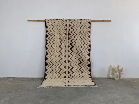 Beige and dark brown zigzag patterned moroccan handmade beni ouarain rug with fringed edges hanging on wooden rod