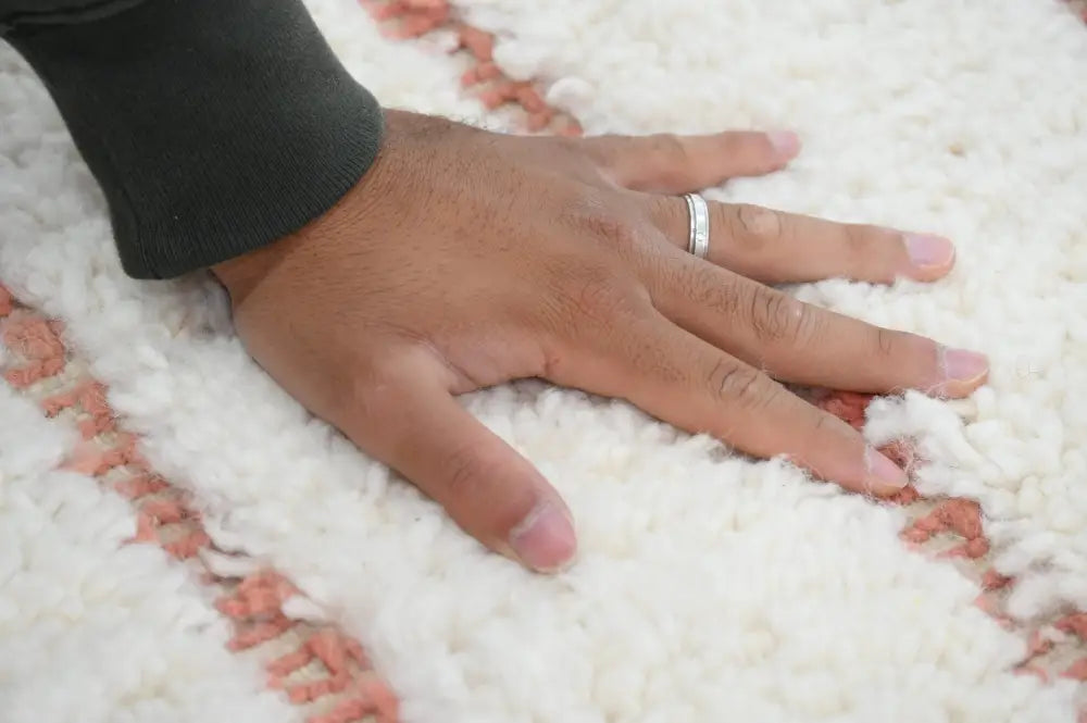 A hand rests on a soft white moroccan handmade beni ouarain rug with subtle pink woven stripes and plush texture