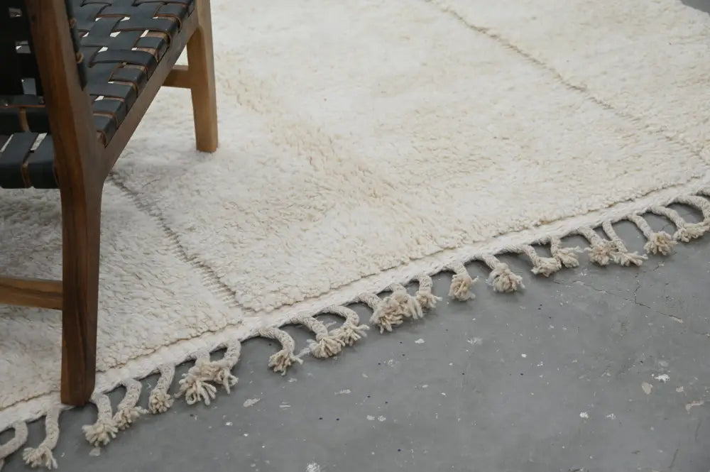 Cream-colored plush wool beni ouarain rug with fringed edge and woven leather seat of chair partially visible