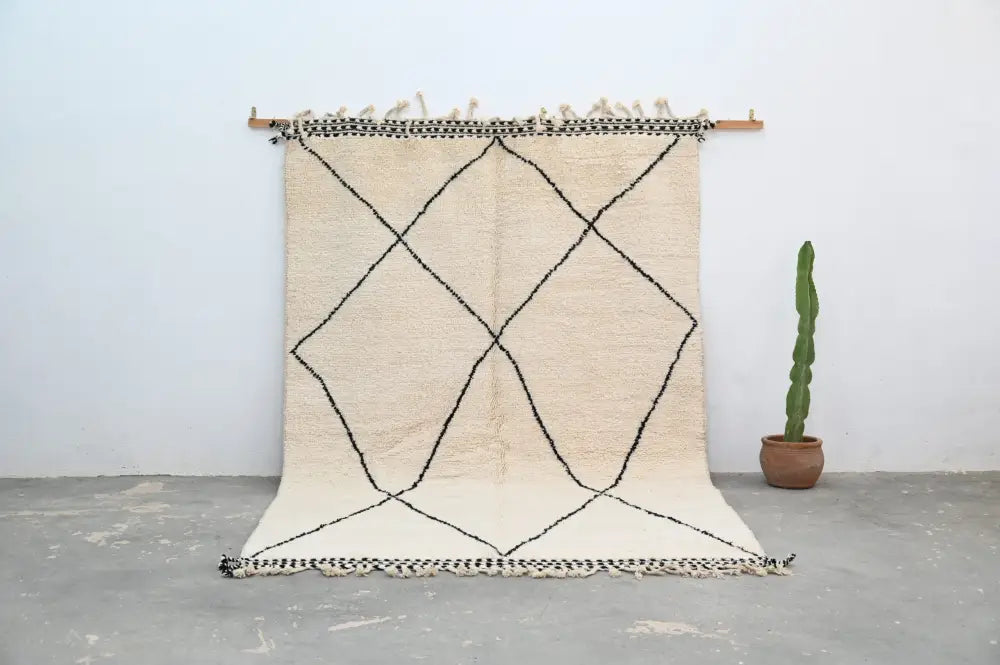 Cream-colored beni ouarain rug with black geometric diamond patterns and fringed edges hanging on wooden rod