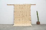 Cream-colored woven decorbites™ handmade traditional azilal rug with zigzag pattern and fringed edges on wooden rod