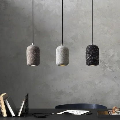 Three cylindrical cement pendant lamps in gray white and black with speckled finish hang against a textured wall industrial style