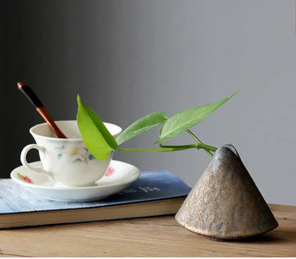 A rustic cone-shaped ceramic pottery vase ornament with metallic gold and gray finish holding a green leafy stem for tea ceremony