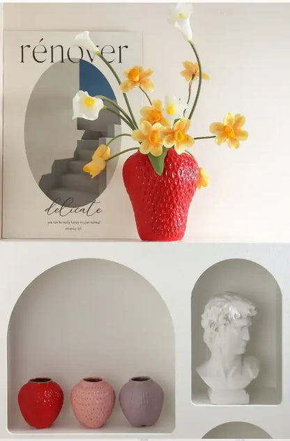 A vibrant red ceramic strawberry cartoon flower vase holding yellow and white calla lilies by DecorBites