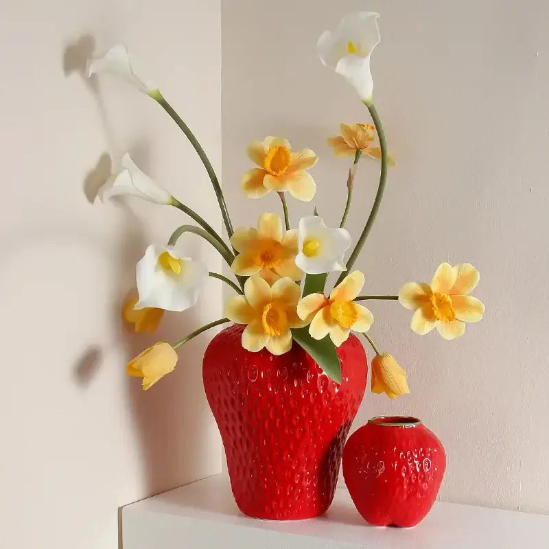 A vibrant red ceramic flower vase with a textured cartoon strawberry surface holding white calla lilies and yellow daffodils