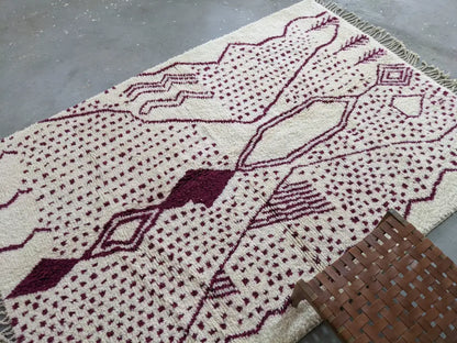 Cream-colored tapis beni ouarain rug with deep burgundy geometric patterns and fringed edges by DecorBites™