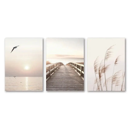 Set of three minimalist white seascape canvas prints featuring a seagull, boardwalk, and tall reeds by DecorBites™