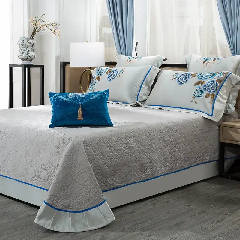 Light gray quilted bedspread with blue piping and ruffled edges in an elegant ivory and royal blue 200s embroidery egyptian cotton bedding set