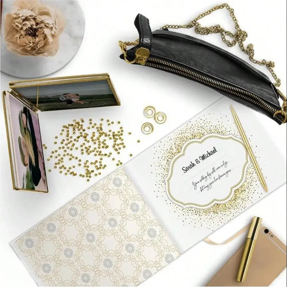 Black leather crossbody pouch with gold-tone zipper and chain strap alongside elegant white decorbites guest book with bronzing detail