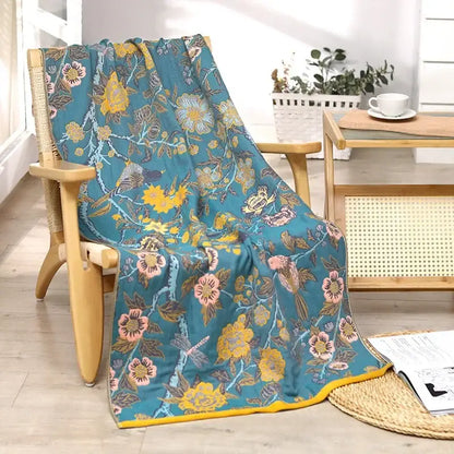 A teal floral bath towel with yellow and pink blossoms birds and dragonflies made of decorbites cotton gauze fabric draped on a chair