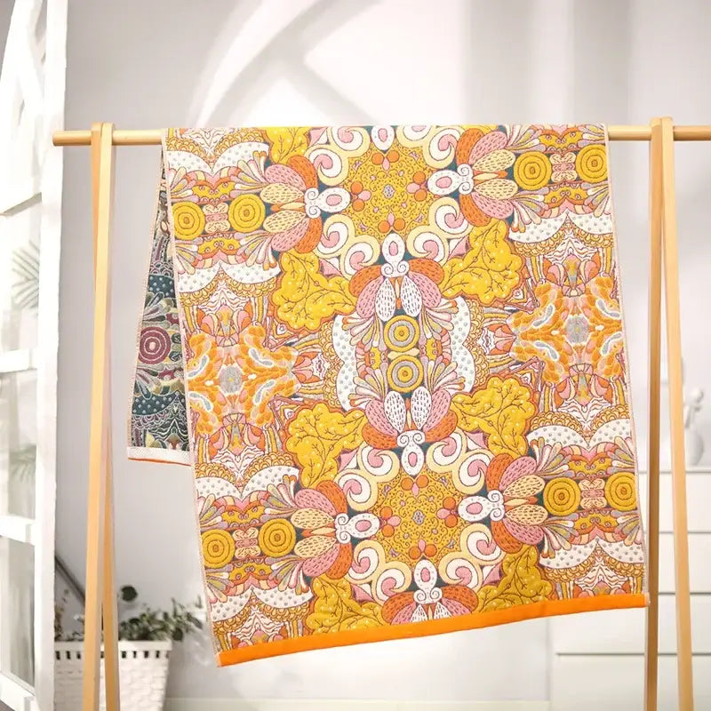 A vibrant yellow and pink floral patterned cotton gauze bath towel with bold orange border hanging on wooden rack decorbites cotton gauze bath