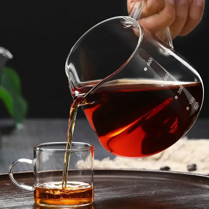 Fluted clear glass coffee pot with measurement markings pouring dark amber tea into matching glass coffee cup