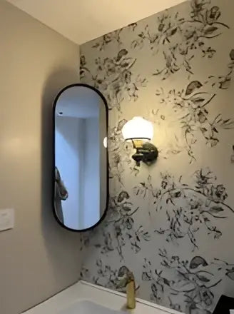 French retro brass and black wall lamp installed in a bathroom with floral wallpaper and an oval mirror above a bathtub