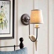 A cream ceramic bird decor antique wall lamp with a bronze arm and beige linen shade for bedroom decorbites wall lamp