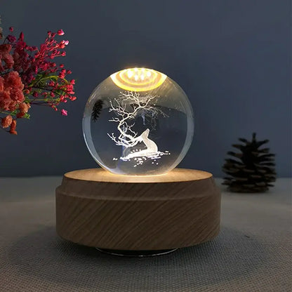 A clear glass crystal ball music box with an etched white deer design and warm yellow light on a wooden base