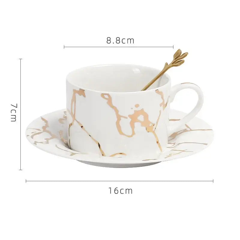 White ceramic cup and saucer set with gilded marble texture and matching golden leaf-shaped spoon by decorbites