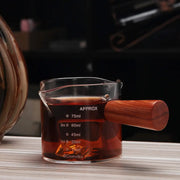 Glass espresso pitcher with clear glass body and polished wooden handle marked in milliliters and ounces