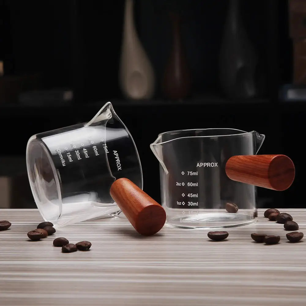 Clear glass espresso pitcher with wooden handle and measurement markings for precise kitchen use