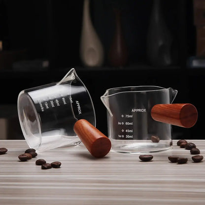 Clear glass espresso pitcher with wooden handle and measurement markings for precise kitchen use