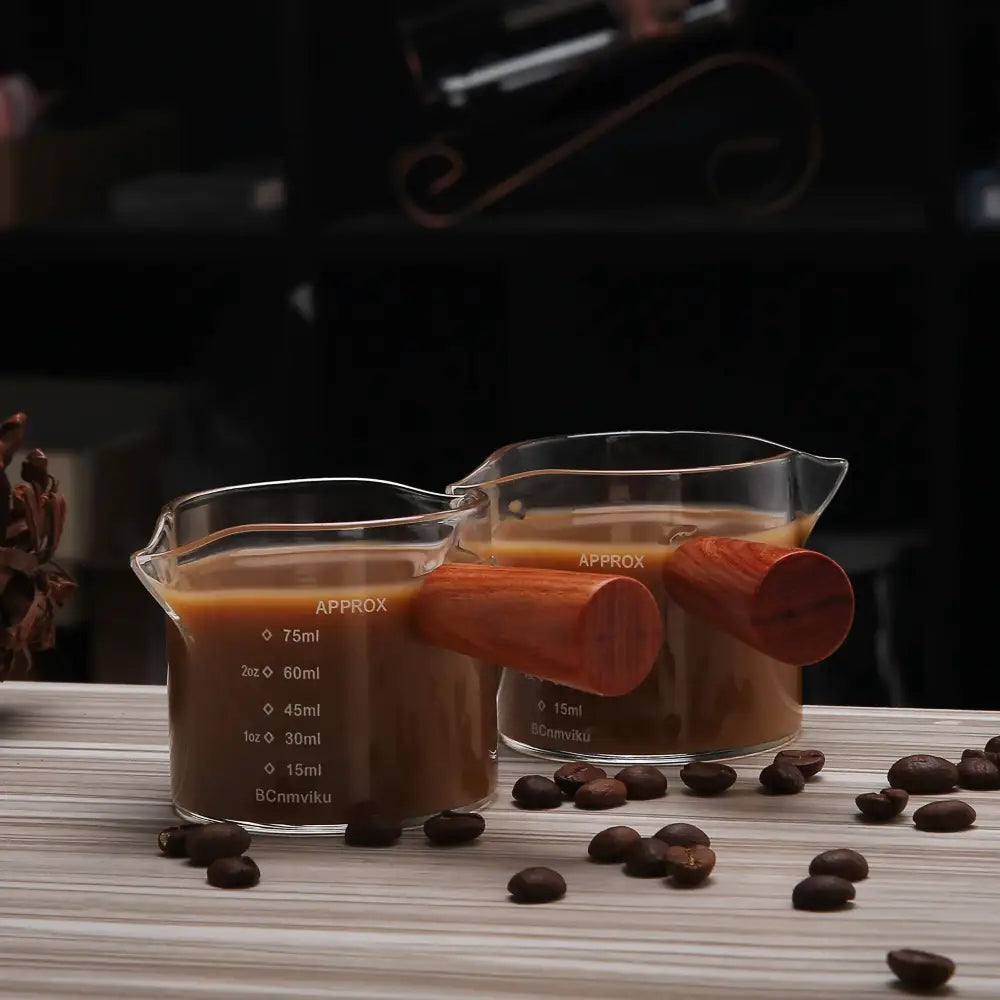 Two clear glass espresso pitchers with wooden handles filled with coffee and marked with volume measurements