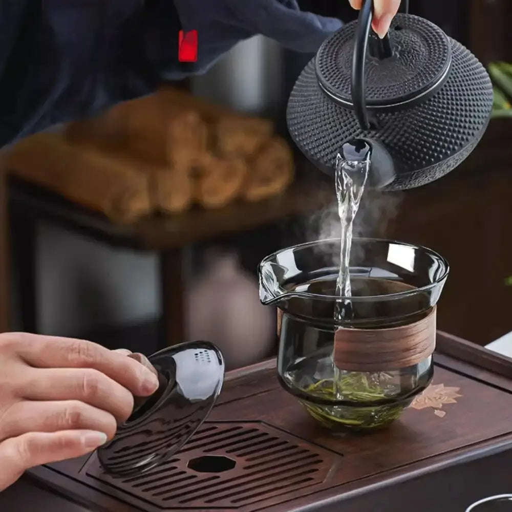 Black cast iron teapot pouring steaming water into a glass gaiwan cup set with wood ring tea filter