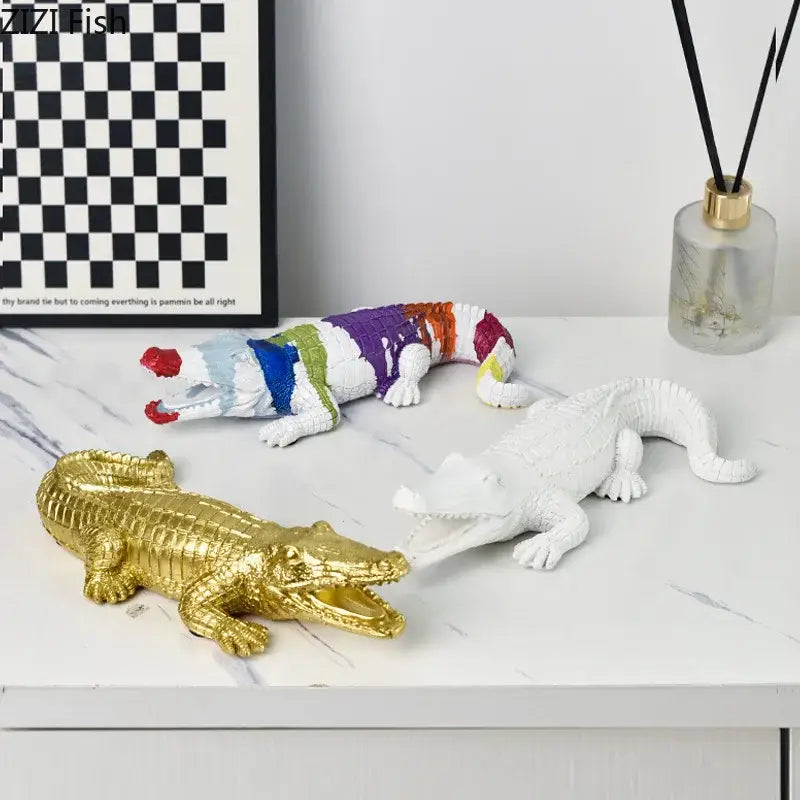 Three decorative decorbites crocodile resin sculptures in gold metallic, white plastic, and vibrant rainbow stripes for desk decor