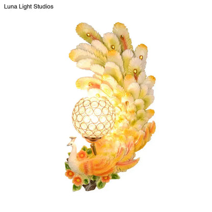 Golden peacock wall light with crystal shade featuring yellow green feathers and a glowing spherical bulb for living room use