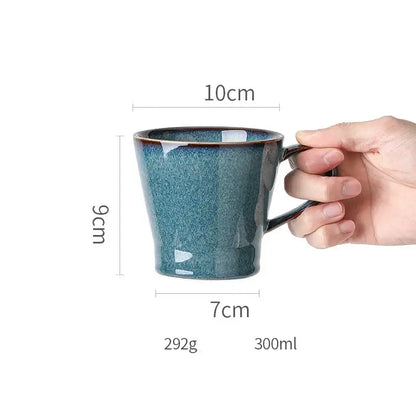 A hand holds a beautiful blue ceramic mug with speckled teal glaze and dark brown rim, complementing dinnerware dish