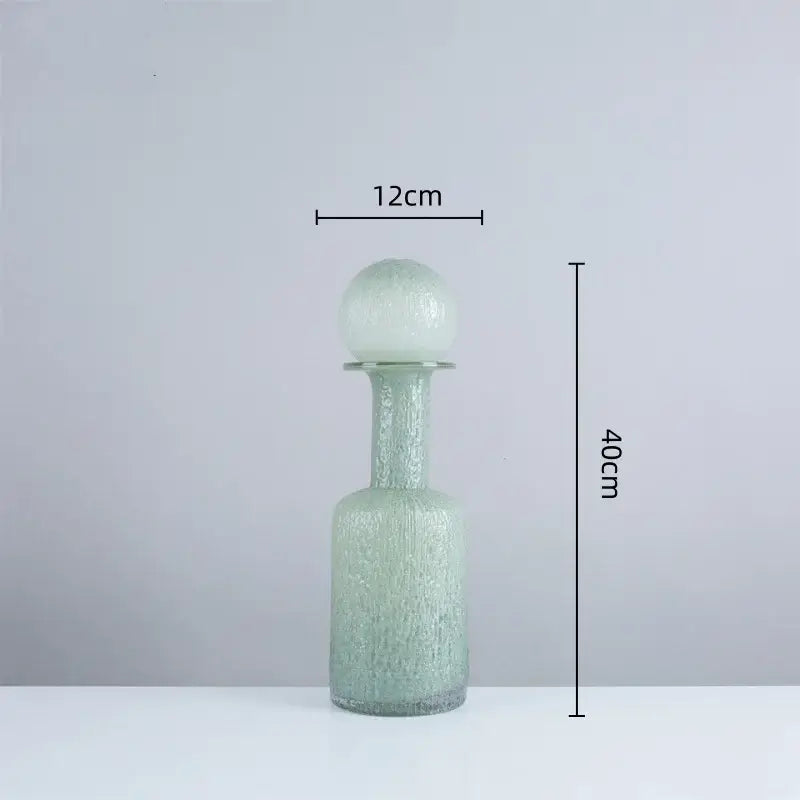 A tall translucent green glass decanter with textured surface and spherical stopper in the decorbites grey round glass vase set with hammer lid for living room decor