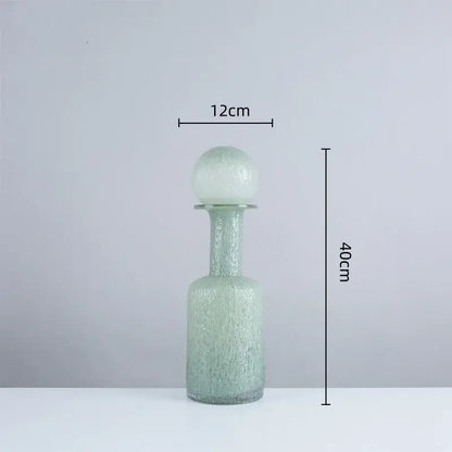 A tall translucent green glass decanter with textured surface and spherical stopper in the decorbites grey round glass vase set with hammer lid for living room decor