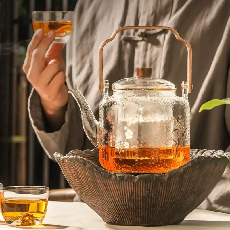 Clear glass teapot with delicate floral etching and copper-colored handle filled with amber tea by decorbites glass teapot