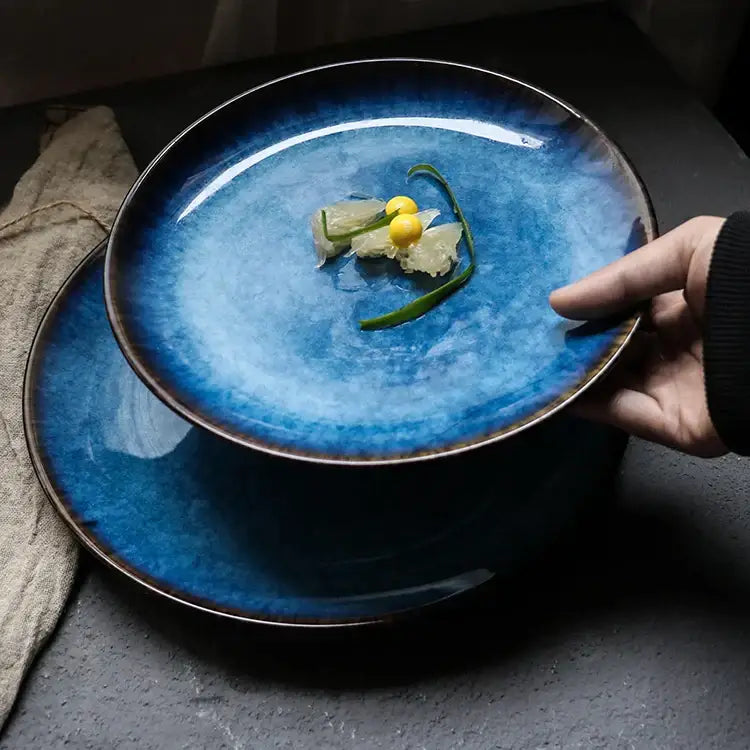 A hand holds a handcrafted ceramic round plate with deep blue gradient glaze and dark brown rim from a dining set of 3
