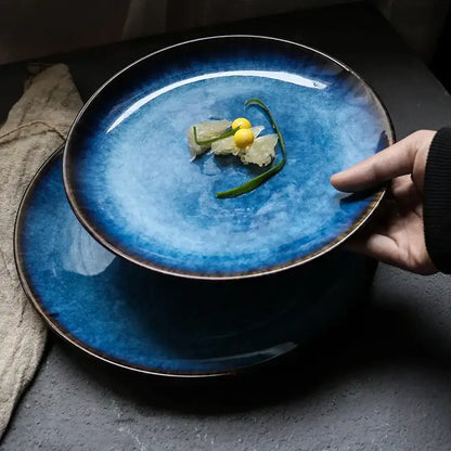 A hand holds a handcrafted ceramic round plate with deep blue gradient glaze and dark brown rim from a dining set of 3