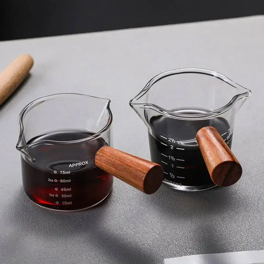Two clear heat resistant glass espresso milk jugs with wooden handles filled with dark liquid and measurement indicators