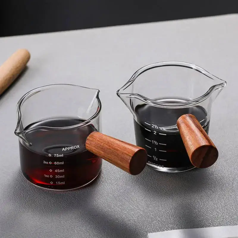 Two clear heat resistant glass espresso milk jugs with wooden handles filled with dark liquid and measurement indicators