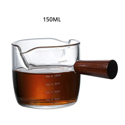 Heat resistant glass espresso milk jug with dark wood handle filled with amber liquid marked for 150ml capacity