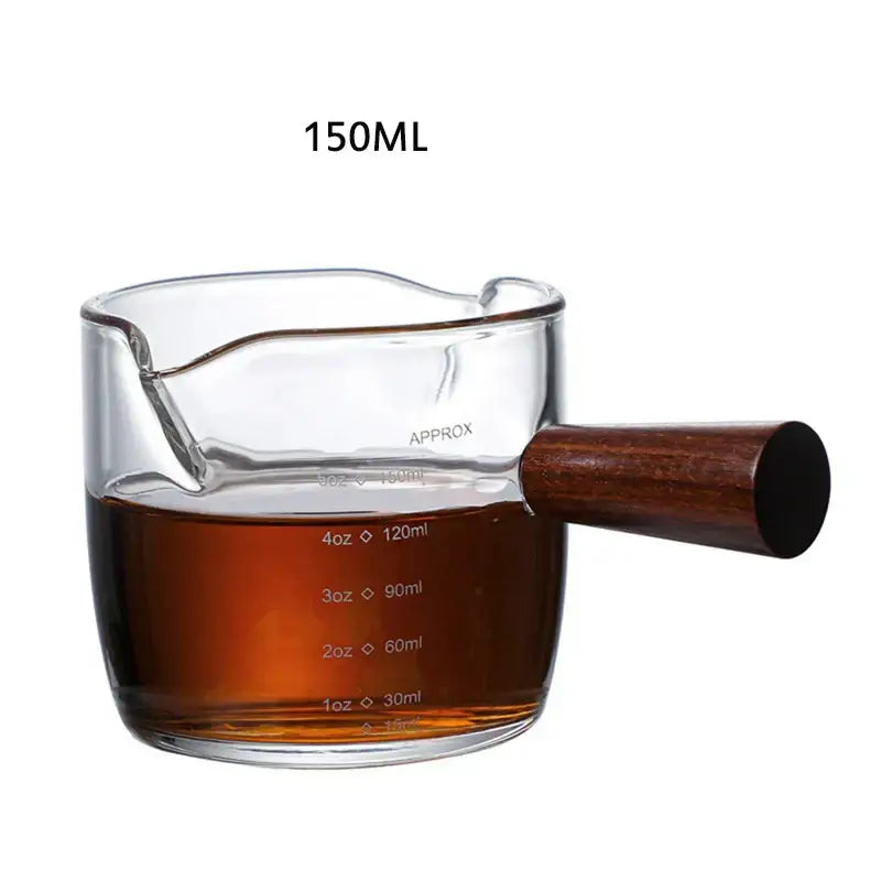 Heat resistant glass espresso milk jug with dark wood handle filled with amber liquid marked for 150ml capacity