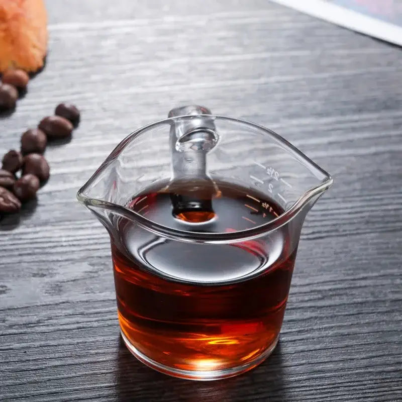 A heat resistant glass espresso milk jug with wood handle filled with dark amber liquid and measurement markings
