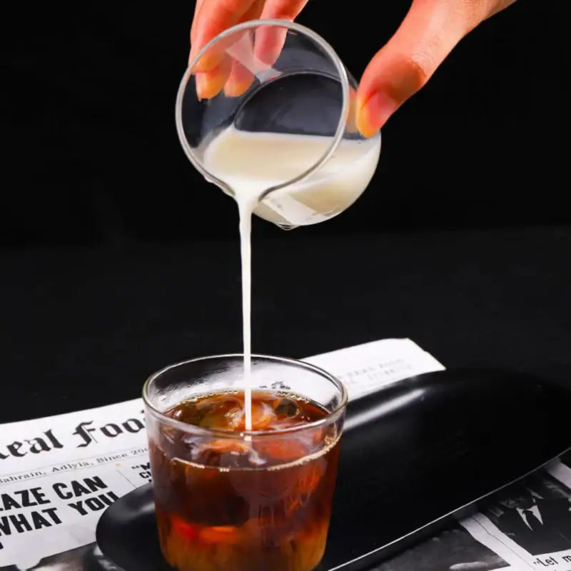 A clear heat resistant glass espresso milk jug with wood handle pouring creamy milk into dark iced coffee