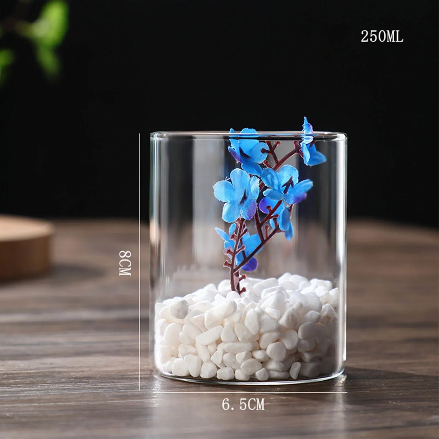 Clear glass cylindrical hydroponics planter vase filled with white pebbles and blue artificial flowers for desktop decor