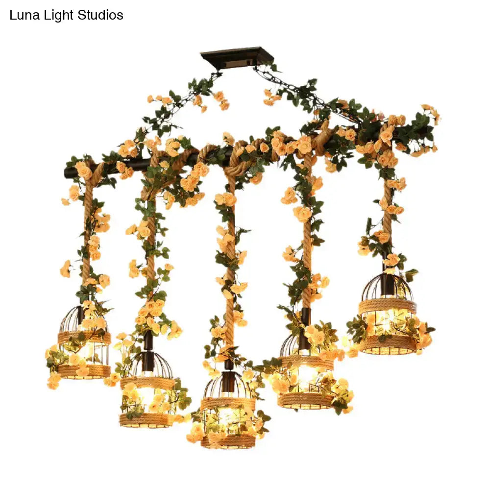 A rustic black decorbites™ birdcage chandelier with five lights wrapped in industrial hemp rope and adorned with yellow roses for restaurant island lighting