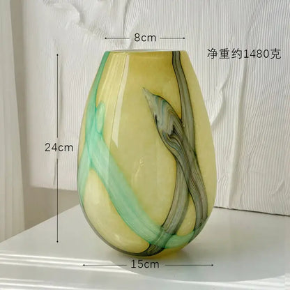 A glossy teardrop-shaped glass vase with irregular geometric green and dark gray marbled patterns on a pale yellow base decorbites irregular geometric decor
