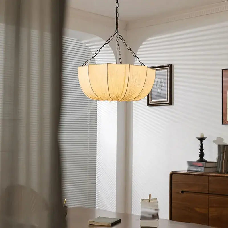 A warm cream-colored pendant light with a pleated white fabric shade and black chain, a refined choice for hospitality settings
