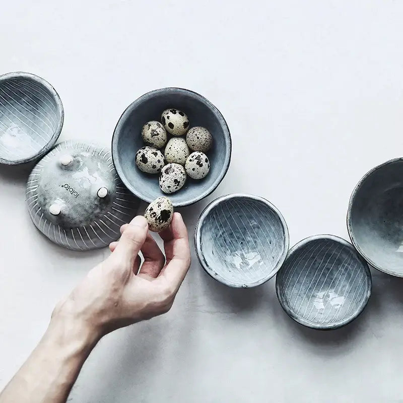 A hand holds a speckled quail egg above Japanese style matte gray small sauce dishes rice bowl with vertical ridges
