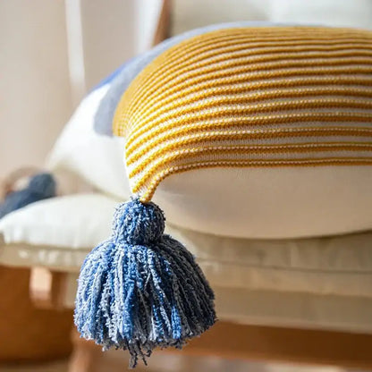 A cozy mustard-yellow and cream decorbites knit stripe pillow cover with a fluffy blue tassel for living room decor